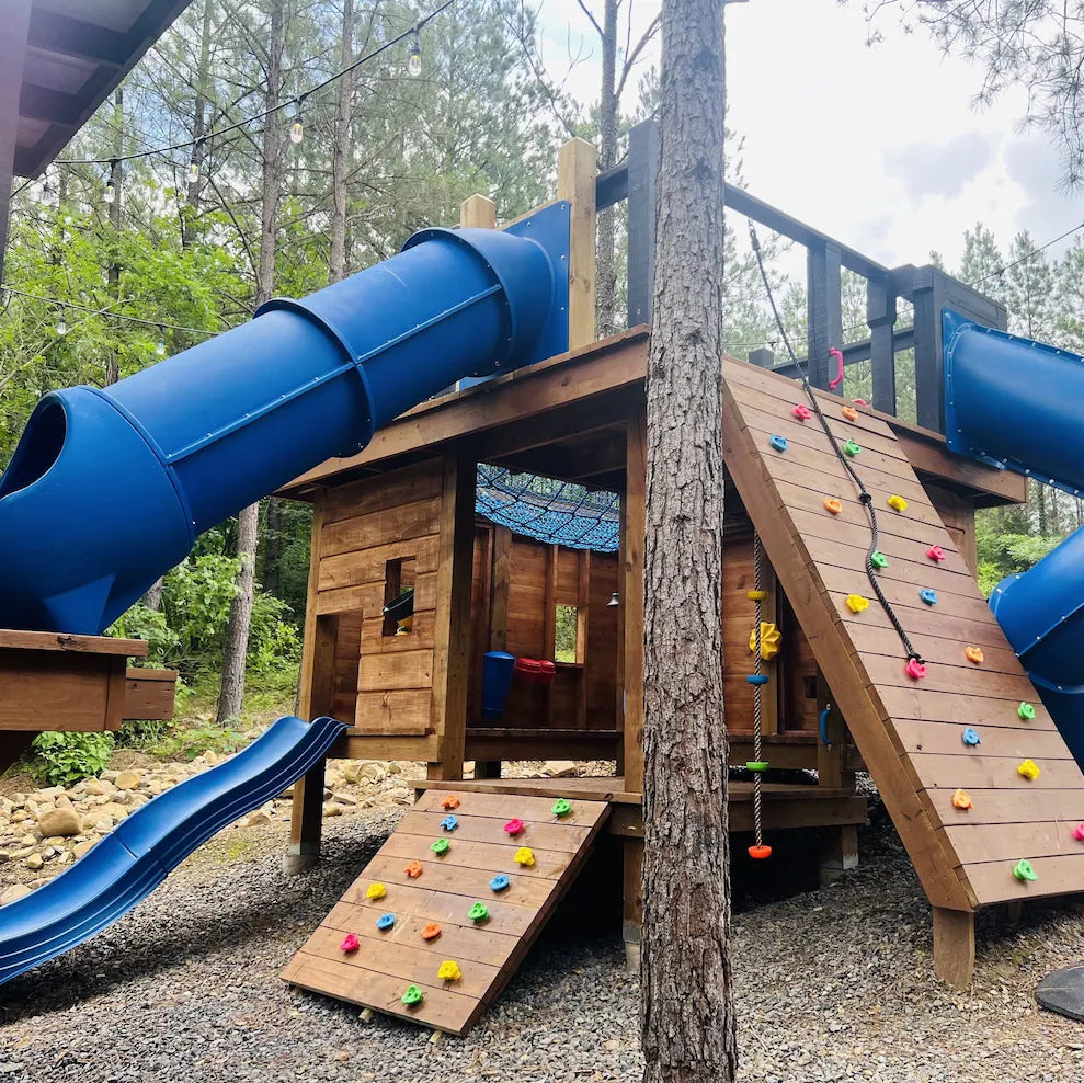 Children's playground with wooden playhouse, blue slides, and climbing wall in a forest setting.