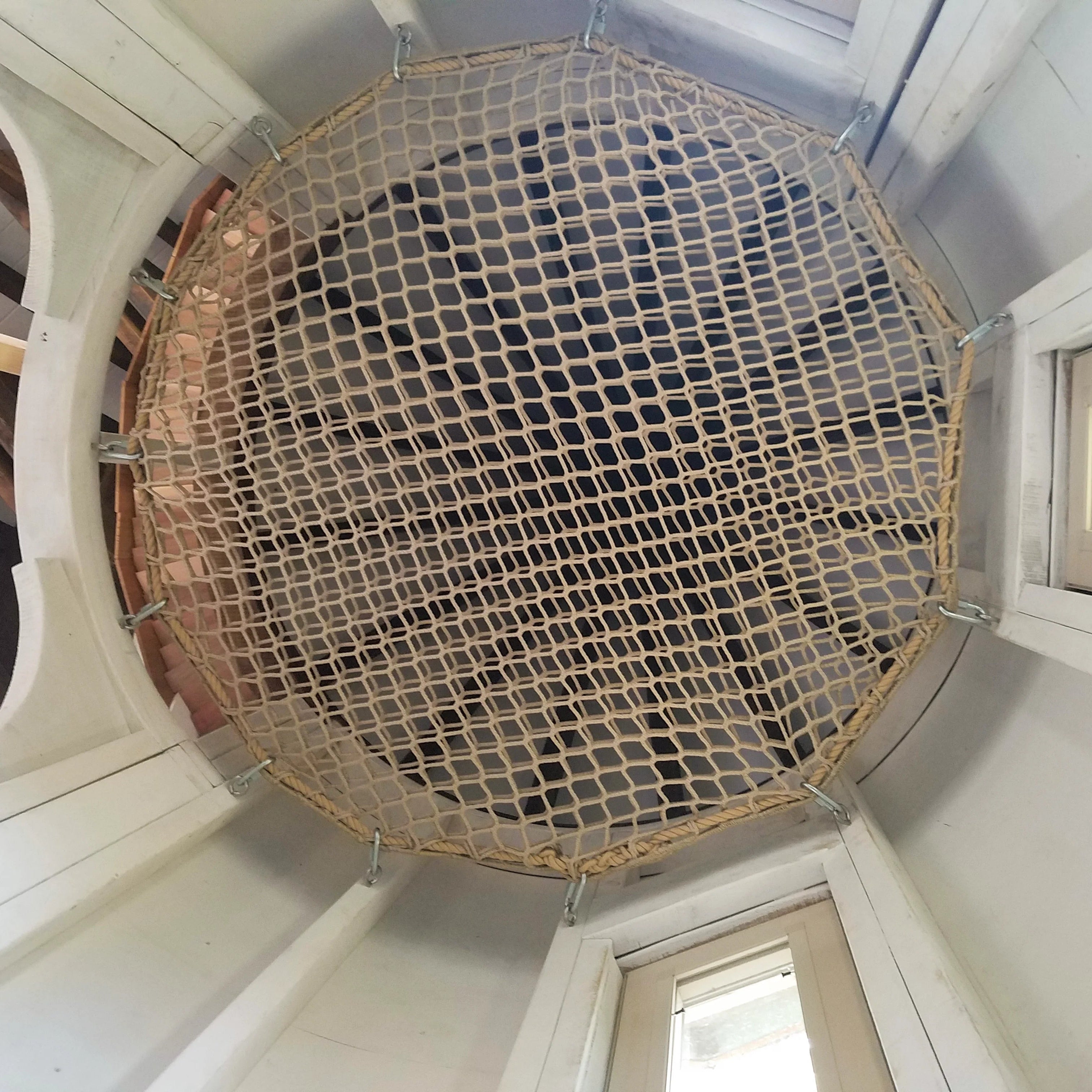Round woven loft hammock net suspended from ceiling in a room with wooden beams and a window.