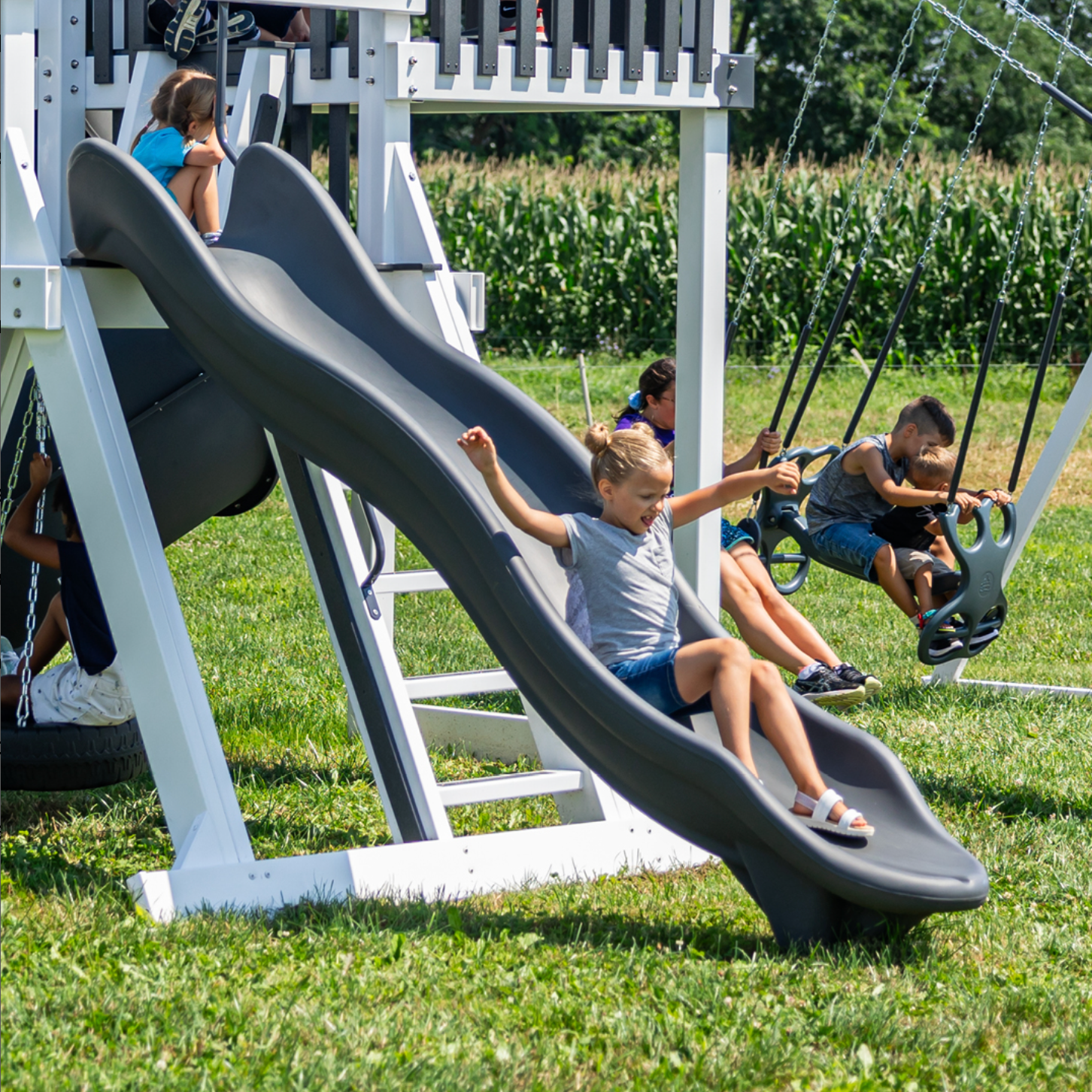 Children playing on a playground slide and swings in an outdoor setting with cornfields in the background.
