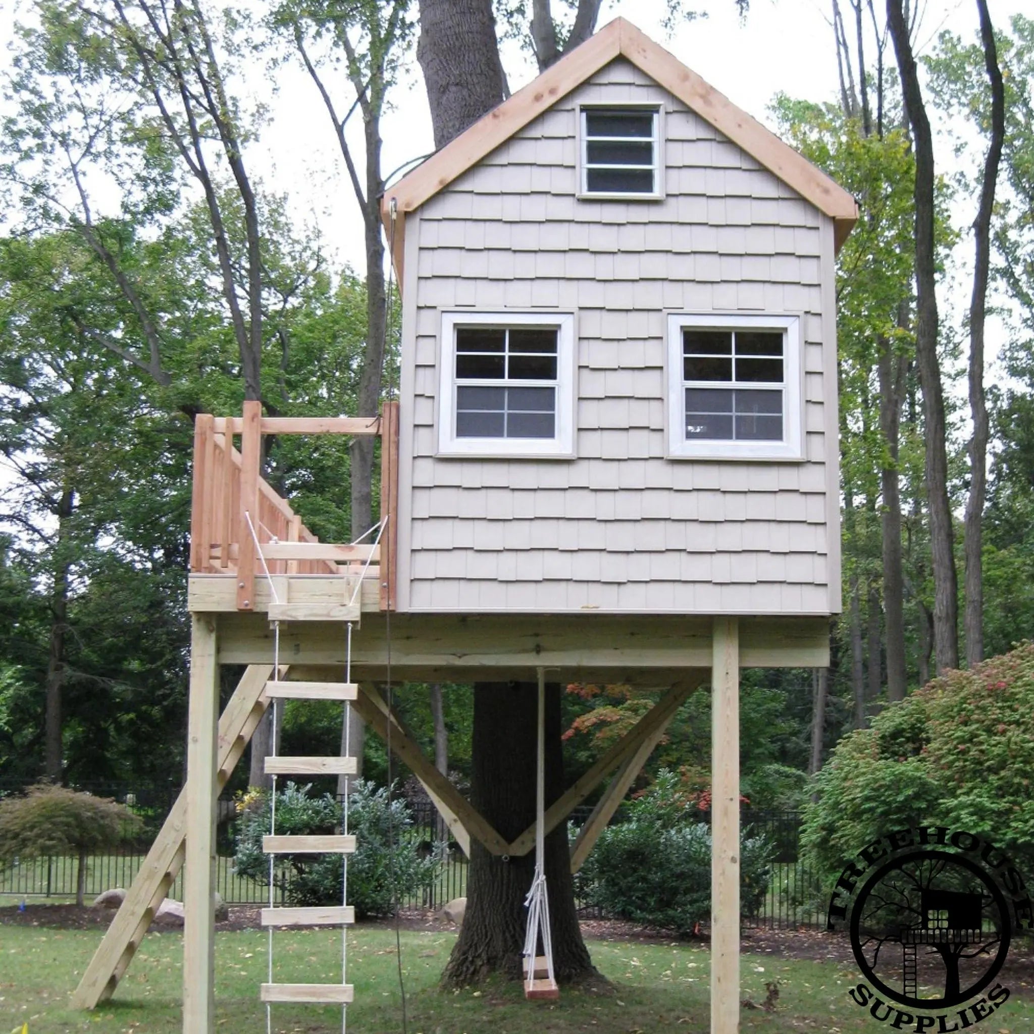 White treehouse 12 foot by 14 foot with rectangular design and a One tree two post layout. Treehouse with deck, white siding, three windows, rope ladder, brown railing and tan roof. Treehouse plans for sale.